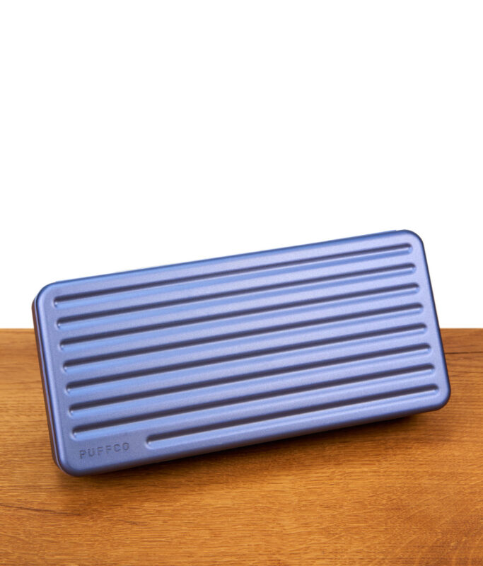 Das geschlossene Travel Case Sapphire Edition horizontal auf einem Holztisch platziert; elegantes saphirblaues Hardcase mit geprägtem Puffco-Schriftzug für den sicheren Transport unterwegs.