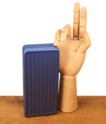 Geschlossenes Puffco Travel Case Sapphire in vertikaler Ansicht neben einer dekorativen Holzhand; gut erkennbar ist die geriffelte, robuste Oberfläche des saphirblauen Etuis.