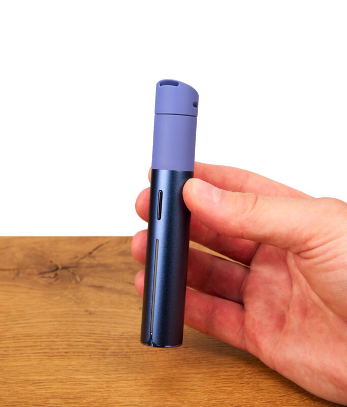 Eine Hand hält den geschlossenen Puffco Pivot Sapphire Edition Vaporizer-Pen. Das Bild zeigt das elegante, mattblaue Saphir-Design und die ergonomische Form des tragbaren Dab-Pens in voller Länge vor einem Holzuntergru