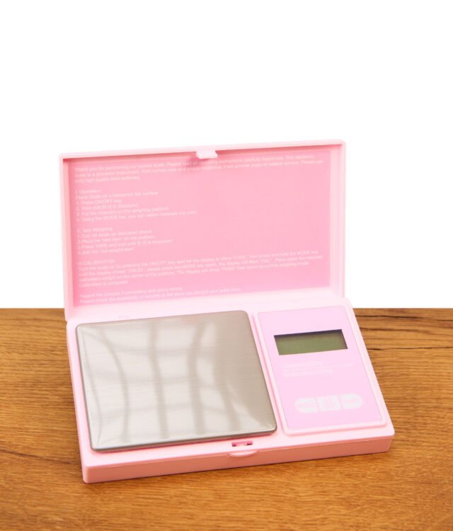 Die geöffnete rosa Glossy Genetics Digitalwaage 0,01-500g Taschenwaage steht auf einer Holzoberfläche und zeigt die metallische Wiegefläche sowie die englische Kurzanleitung im Deckel.