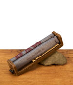 RAW Eco 2-Way Roller Drehmaschine King Size 110mm aus Hanfplastik mit bedrucktem Rollband, gestützt auf einen Stein auf einem Holztisch.
