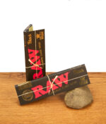 Zwei Päckchen RAW Black King Size Wide Paper. Die schwarzen Heftchen mit rotem Logo und Goldprägung liegen auf Holz und Stein.
