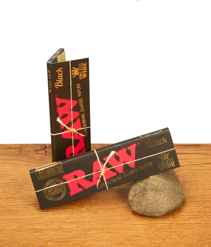 Zwei Päckchen RAW Black King Size Wide Paper. Die schwarzen Heftchen mit rotem Logo und Goldprägung liegen auf Holz und Stein.