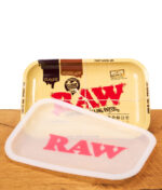 RAW Dab Rolling Tray aus Metall neben dem zugehörigen abnehmbaren Silikonschutz mit pinkem Logo auf einer Holzfläche.