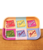 Smoking Rolling Tray Artist Collection aus Metall mit sechs bunten Retro-Motiven von Smoking-Blättchen im Pop-Art-Stil auf einem Holztisch.