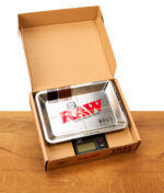 Geöffnete Verpackung der RAW Tray Scale Waage mit schwarzer Digitalwaage und Metall-Tray auf einem Holztisch.
