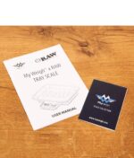 Gedruckte Bedienungsanleitung und Info-Booklet der digitalen RAW Tray Scale Waage auf einem Holztisch.
