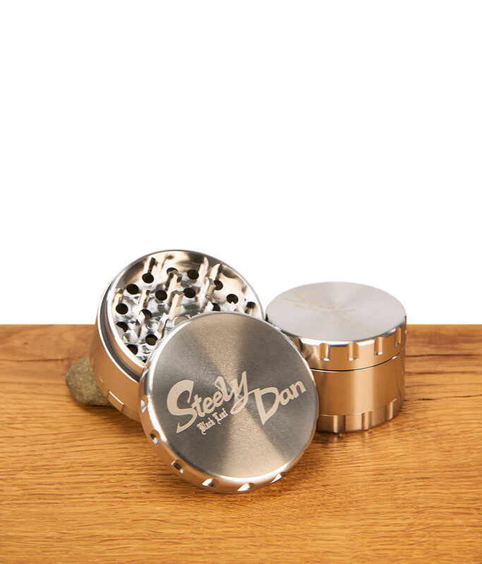 Geöffneter Steely Dan 2.0 Edelstahl Grinder mit CNC-gefrästen Zähnen und graviertem Deckel auf einem Holztisch.