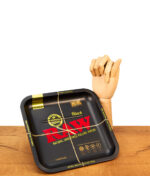 Quadratisches RAW Square Rolling Tray Black aus Metall. Die schwarze Drehunterlage lehnt neben einer Holzhand auf einem Holztisch.