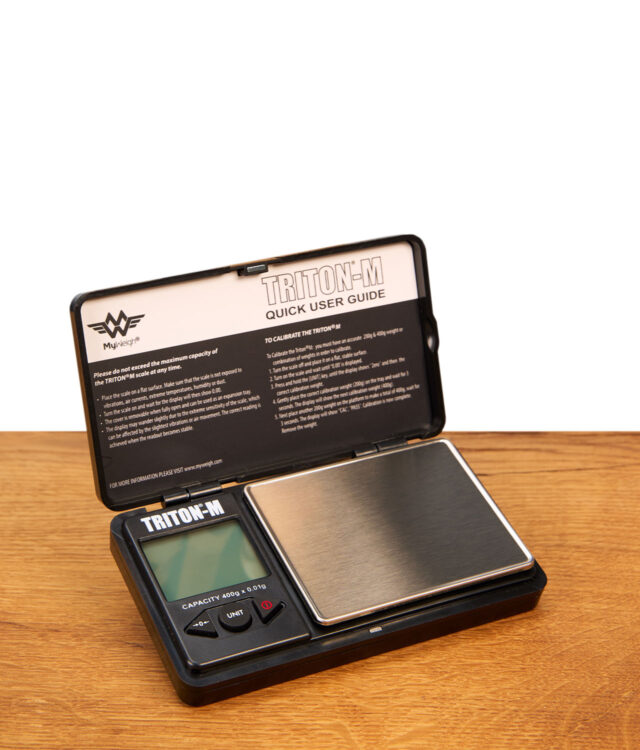 Geöffnete MyWeigh Triton-M digitale Taschenwaage mit Edelstahl-Wiegefläche und Schutzdeckel auf Holzfläche.