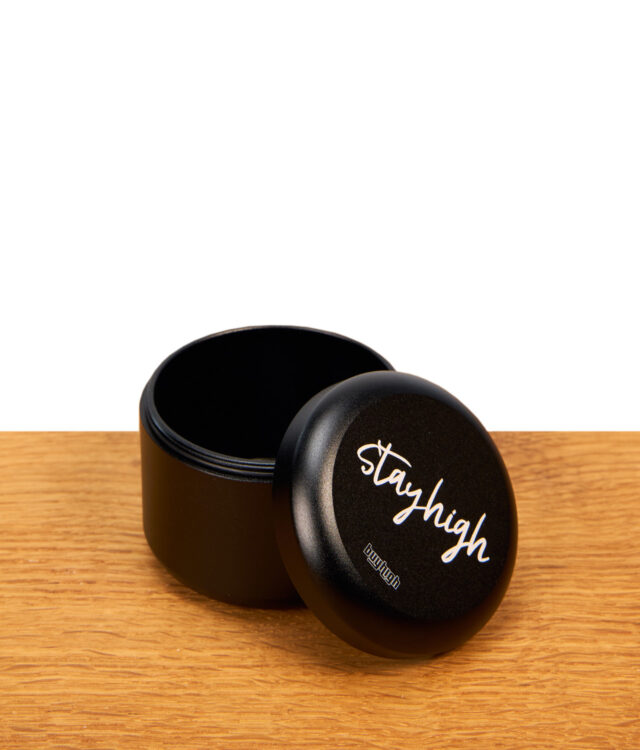 Geöffnete Stayhigh Puck Large Kräuterdose aus mattschwarzem Aluminium mit danebengelehntem Deckel auf Holz.