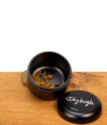 Geöffneter Stayhigh Puck Large Stash-Behälter aus Aluminium mit getrockneten Kräutern auf einer Holzoberfläche.