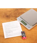 Silberne Black Leaf Feinwaage mit zwei AAA-Batterien und der Bedienungsanleitung auf einer hölzernen Arbeitsplatte.