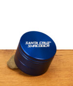 Dunkelblauer Santa Cruz Shredder grinder in 3-teiliger ausführung mit weißem Logo steht angewinkelt auf einem Stein, präsentiert auf Holz.