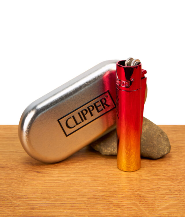 Das schimmernde Clipper Metall Sunset Gradient Feuerzeug steht aufrecht neben seiner geschlossenen, ovalen Präsentationsbox aus Metall auf einem Holztisch vor weißem Hintergrund.