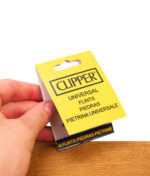 Gelbe Blisterverpackung der Clipper Universal Feuersteine (9er Pack) in einer Hand gehalten über Holz.