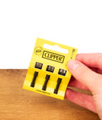Hand hält das verpackte Clipper Flintsystem 3er Pack mit Ersatz-Zündstiften und Metallrädchen über einen Holztisch.