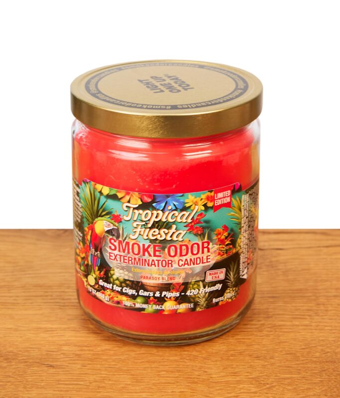 Geschlossene Smoke Odor Duftkerze Tropical Fiesta im Glas auf Holz. Rotes Wachs und buntes Tropen-Etikett unter goldfarbenem Metalldeckel.