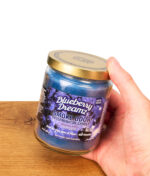 Hand hält die geschlossene Smoke Odor Duftkerze Blueberry Dreamz. Glasgefäß der Anti-Geruch Kerze mit blauem Wachs.