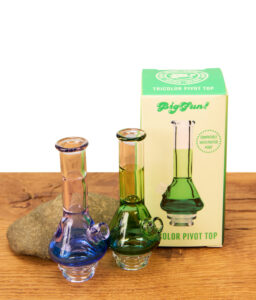 Blauer und grüner BigFun! Tricolor Pivot Top Glasaufsatz neben der grünen Originalverpackung und Stein auf Holz.