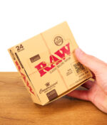 Hand hält eine geschlossene RAW Connoisseur King Size Slim Papers Displaybox aus Karton auf Holz.