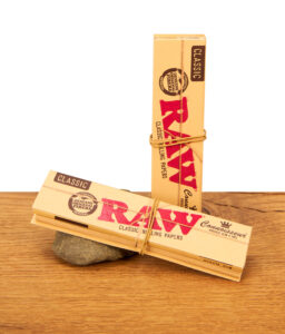 Zwei RAW Connoisseur King Size Slim Heftchen mit Tips und Gummiband dekorativ auf Holz und Stein.