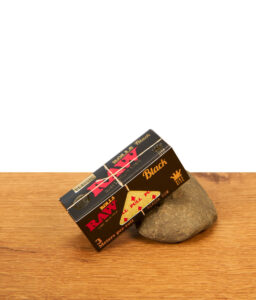 RAW Black King Size Roll Schachtel aus Karton gelehnt an einen Stein auf Holzuntergrund.